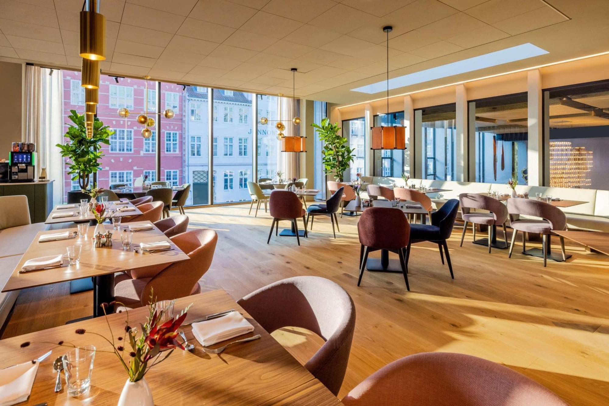 Nh Collection Copenhagen Hotel Kültér fotó The photo depicts a modern restaurant interior with ample natural light. The space features large windows that reveal buildings outside, enhancing the ambiance. Inside, there are several dining tables arranged with elegant table settings, including c